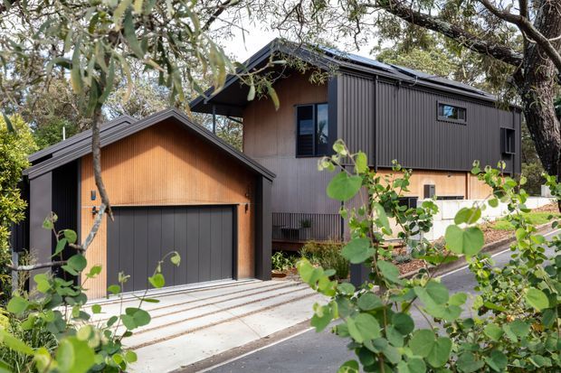 Exterior view of a contemporary two-storey dwelling in a leafy setting, featuring dark vertically ribbed metal cladding, warm timber wall panels, a pitched roof, and a detached single-car garage with a broad dark garage door. A concrete driveway and landscaped planting occupy the foreground.