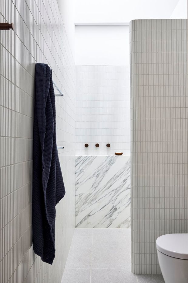 A bright, narrow contemporary bathroom with vertically ribbed light tile walls, pale floor tiles, and a curved tiled partition screening a white toilet. At the rear is a marble-look tiled wall with three dark circular wall fixtures and a small basin or shelf. A dark navy towel hangs from a wall-mounted rail, adding contrast to the otherwise white and grey palette.