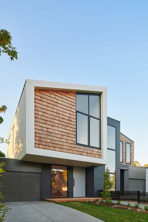 Exterior view of a contemporary multi-level residence with a projecting upper-storey volume. The facade combines dark horizontal cladding, warm terracotta-toned shingle cladding, crisp white framing, and large dark-framed windows. A concrete driveway, landscaped garden, entry door, and black metal fence are visible under a clear blue sky.