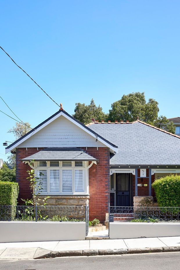 Street-facing photograph of a compact heritage-style brick bungalow with a prominent front gable, slate-clad pitched roof, white weatherboard gable detailing, timber-framed windows with shutters, a covered entry, decorative wrought-iron fencing, and a low rendered front wall. Mature trees and neighbouring homes are visible behind the property.