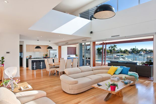 Bright open-plan contemporary living area with a large curved sectional sofa, timber flooring, a glass-topped coffee table, and a modern kitchen with island seating. Floor-to-ceiling glazing opens toward a waterfront view, while a dramatic skylight and double-height ceiling bring in abundant daylight. Sculptural pendant lights and colourful cushions add accents.