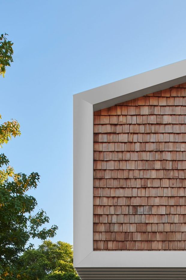 Close-up exterior architectural detail showing a contemporary building corner with a thick white rendered frame around a wall clad in small overlapping reddish-brown timber shingles. A clean sloping roofline and layered soffit are visible, with leafy trees and clear blue sky in the background.
