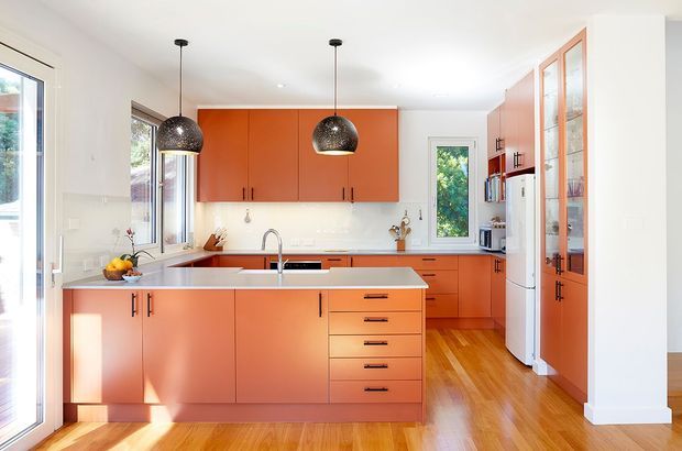 Bright contemporary kitchen with a large central island, vivid orange built-in cabinetry, white benchtops and splashback, timber flooring, tall glazed pantry-style cabinetry, and two black pendant lights. Large windows and a glazed door bring in abundant natural light and views of greenery.