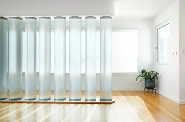 Bright minimalist interior featuring a distinctive freestanding partition of tall translucent cylindrical panels mounted on a curved timber base. The room has white walls, large windows, honey-toned timber flooring, and a potted plant on a small stand. Strong daylight creates a clean, airy atmosphere.