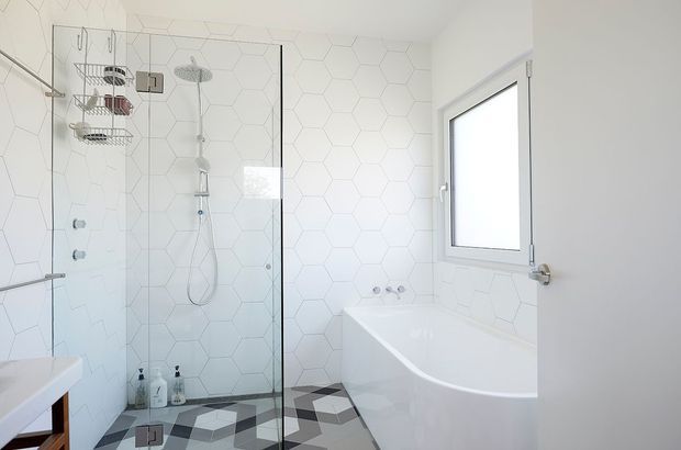 Bright contemporary bathroom featuring a glass-enclosed walk-in shower, freestanding white bathtub, white hexagonal wall tiles, geometric patterned floor tiles, and minimal chrome fixtures. A frosted window provides natural light.