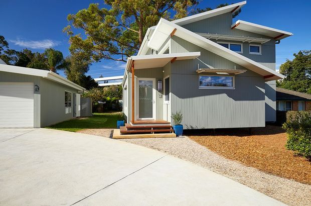 Exterior view of a compact multi-level grey-clad house with distinctive overlapping pitched roof forms, white fascia and trims, a small covered entry porch, timber steps, and adjacent detached garage. The property sits beside a broad concrete driveway with landscaped gravel and mulch areas, mature trees, and neighbouring residential buildings visible around it.