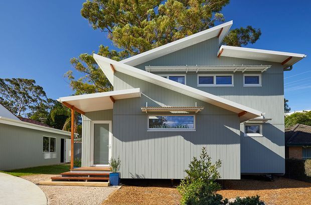 Exterior photograph of a distinctive contemporary two-storey house with overlapping angular roof planes, grey vertical cladding, white fascia and trim, and several windows with external awnings. A small covered entry porch with timber posts and steps is visible at the front, set within a landscaped suburban site.