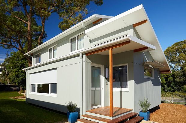 Exterior photograph of a compact contemporary two-storey house with pale grey vertical cladding, crisp white trim, a strongly angled gabled roof form, and a small timber-framed covered entry deck. The facade includes large windows, a glazed entry door, external blinds or awnings, white gutters, and timber soffit and porch details. The home is set within a leafy suburban garden with gravel landscaping.