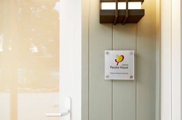 Close-up of a residential exterior entry door and adjacent wall panel, featuring a clearly visible Certified Passive House plaque. The door has a light-coloured frame and handle, while the adjoining vertical-panel surface is muted grey-green with a dark wall-mounted light fitting above the plaque. The image is tightly framed around the entrance and contains no visible interior room.