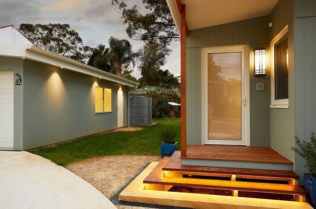 Exterior view of a contemporary single-storey dwelling or extension, focused on a covered entry porch with illuminated timber steps, a glazed front door, and vertical muted green-grey cladding. A detached or adjacent matching structure, concrete driveway, gravel path, lawn, rainwater tank, and mature trees are visible.