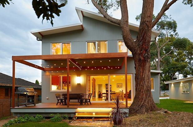 A two-storey contemporary home viewed from the garden at dusk. The grey vertically clad upper level has asymmetrical low-pitched roof planes and large windows, while the ground floor opens onto a timber deck and covered outdoor dining area with warm lighting. A mature tree dominates the foreground, with neighbouring residential structures visible at the sides.