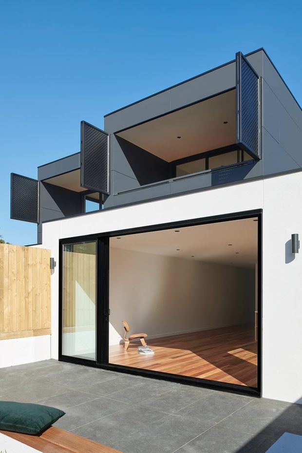 Exterior view of a contemporary multi-level residence with crisp white and charcoal geometric volumes. The upper storey features projecting balconies or recessed terraces enclosed by perforated dark metal privacy screens, while the ground floor opens through large black-framed glazing onto a paved courtyard or patio. Inside, a minimally furnished room with warm timber flooring is visible.