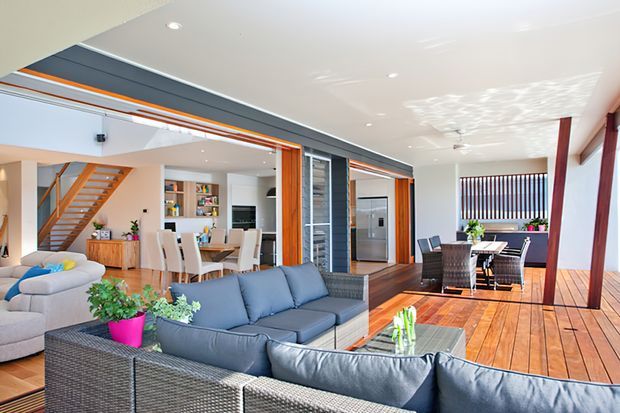 Bright open-plan contemporary home interior combining a living room, dining area and kitchen, with a visible timber staircase, dark feature wall panels, expansive glazing, and a generous timber deck or sunroom extending to the right. The room is furnished with large grey sectional seating and a dining table, with warm timber flooring and orange-toned timber accents.