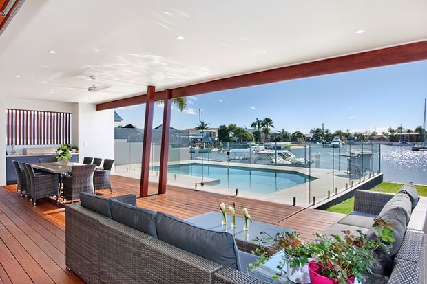 A bright, open-plan waterfront living and dining area opening onto a timber deck and swimming pool. The space features extensive frameless glass balustrading, dark timber structural posts and beams, wicker outdoor-style seating, a long dining table, and views across a canal lined with boats and residences.