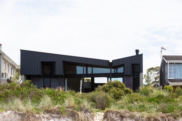 Exterior view of a contemporary, angular single-storey house with dark vertical cladding, extensive horizontal glazing, and a folded roofline. The home sits behind coastal dune vegetation, with neighbouring residential buildings visible on either side.