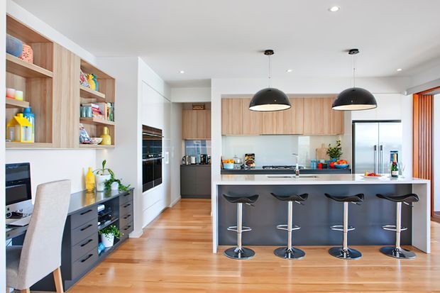 Bright contemporary open-plan kitchen with a large island and four black bar stools, light timber cabinetry, white surfaces, integrated appliances, and black dome pendant lights. A compact home office nook with desk and shelving is visible at the left.