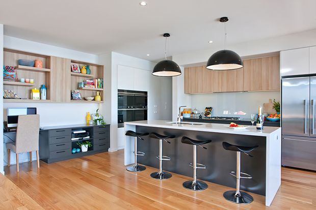 Bright, contemporary open-plan kitchen with a large white island and integrated breakfast bar seating for four. The kitchen features warm timber flooring, light timber upper cabinetry, dark lower cabinetry, a pale splashback, stainless-steel appliances, open display shelving, and black dome pendant lights. A compact work area is integrated into the cabinetry at the left.