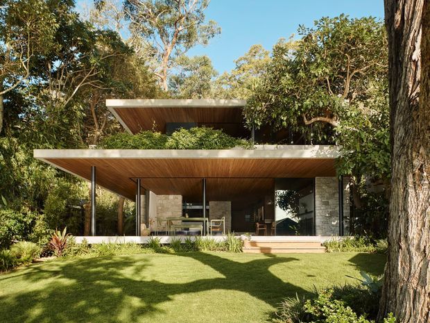 A contemporary pavilion-style house nestled among mature trees and landscaped garden. The low, broad roof planes feature warm timber soffits and deep cantilevered eaves, supported by slender dark posts. Extensive glazing opens onto a lawn and covered outdoor living area, while pale stone masonry forms portions of the walls. The image has a strong indoor-outdoor connection and a calm natural setting.