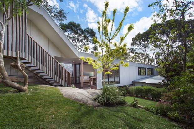 Exterior view of a low-slung contemporary house set within a landscaped, sloping garden. The building features crisp white wall cladding, broad dark-framed windows, a shallow pitched roof, and a prominent timber-railed exterior stair leading to a raised entry or upper level. Mature trees and ornamental planting surround the residence.