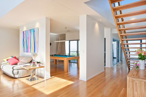 Bright open-plan residential interior with warm timber flooring, white walls, a contemporary living area, a pool table visible through an opening, and an open-riser timber staircase leading upward. Large windows provide natural light, while colourful artwork and cushions add vibrant accents.