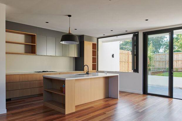 A bright, contemporary open-plan kitchen with a large timber island, integrated cabinetry, open shelving, a pale benchtop, and dark-framed glazing. The kitchen opens to a paved outdoor area through full-height sliding glass doors, while polished timber flooring adds warmth. Recessed ceiling lights and a suspended pendant provide illumination.