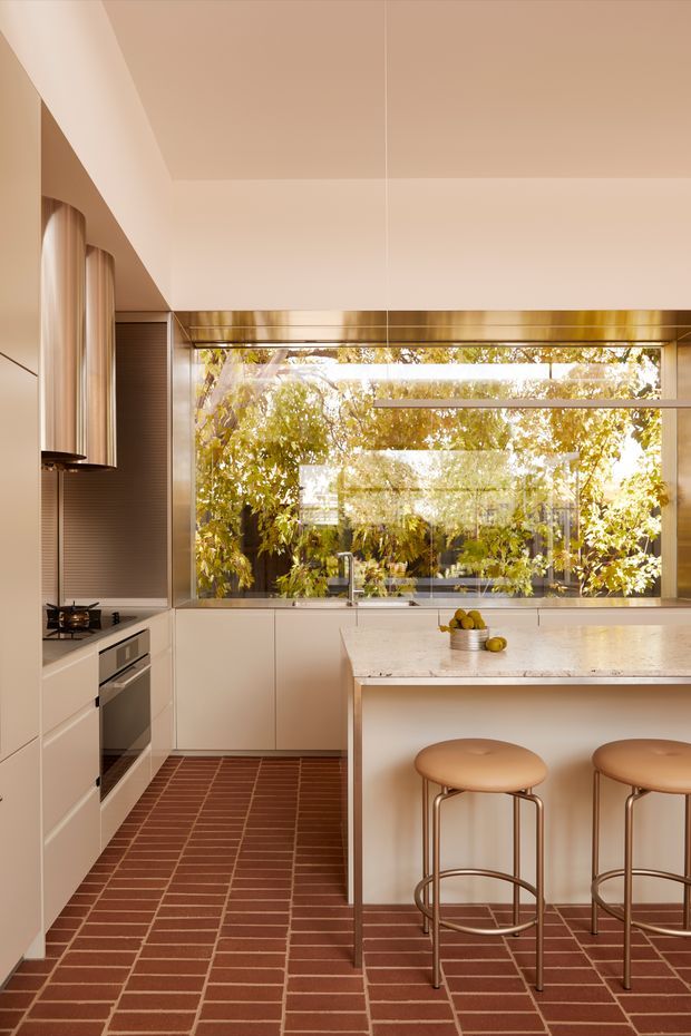 A warm, refined contemporary kitchen with a compact linear layout and central island. The room features pale integrated cabinetry, a large horizontal glazed window overlooking leafy vegetation, a pale stone island benchtop, brass-toned window trim and pendant lighting, and a distinctive red-brown tiled floor laid in a regular grid pattern. Two upholstered bar stools sit at the island.