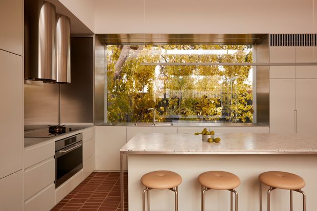 A warm contemporary kitchen with a large central island and three rounded upholstered stools. The space features pale built-in cabinetry, integrated appliances, a stone island benchtop, a dark terracotta-toned tiled floor, and a large glazed opening overlooking leafy trees. Curved metallic-toned upper cabinetry and concealed linear ventilation add refined detailing.