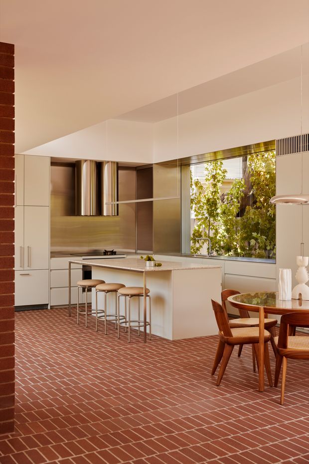 A warm, contemporary open-plan kitchen and dining area. The kitchen features pale integrated cabinetry, a central island with seating for four, a stainless-steel-toned splashback and appliances, and a large window overlooking leafy greenery. The floor is covered in a distinctive reddish-brown brick pattern with light mortar lines. A round glass-topped dining table with timber chairs occupies the foreground. The ceiling is plain and angled, with discreet integrated ventilation and lighting details.