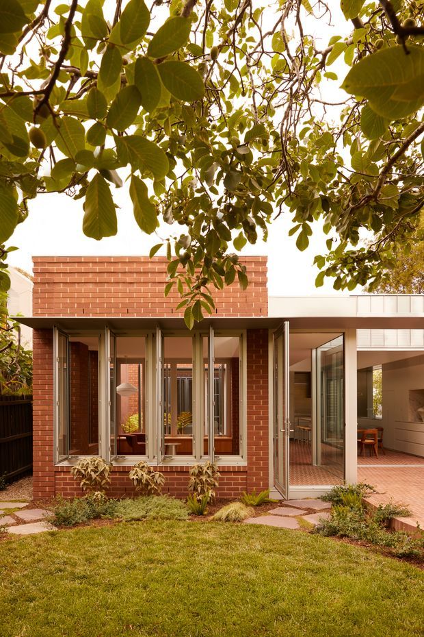 A contemporary residential extension viewed from a landscaped garden. The building features red brick walls, extensive floor-to-ceiling glazed doors and windows, a light-coloured flat roof or parapet, and a covered outdoor terrace leading into the interior. A lawn, stepping-stone path, and mature leafy tree frame the foreground and upper portion of the image.