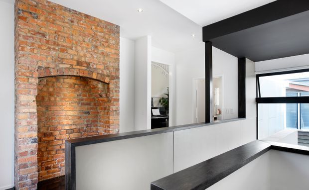 Bright contemporary interior featuring a retained exposed red-brick fireplace and chimney breast on the left, contrasted with crisp white walls and cabinetry. A dark charcoal ceiling beam and structural framing define the open-plan space, while a dark stone or composite benchtop forms a long counter in the foreground. Glazing and a modern neighboring structure are visible at the rear.