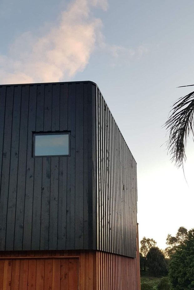 Exterior view of a compact modern building with a tall, simple rectangular form and low-pitched or flat roofline. The upper level is clad in dark vertical boards with narrow ribbed detailing, while the lower level uses warm natural timber cladding. A small horizontal window is set into the dark facade. Trees, palm fronds, and an evening sky surround the structure.
