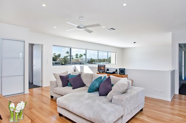 Bright contemporary open-plan living room with a large cream sectional sofa, colourful accent cushions, timber flooring, white walls, recessed lighting and a ceiling fan. Wide windows provide abundant daylight and views toward palm trees and neighbouring buildings. A compact media console and speakers sit along the rear wall, with a partially visible hallway and adjoining spaces.