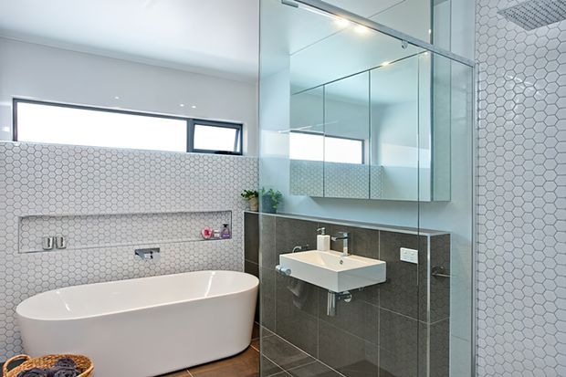 Bright contemporary bathroom featuring a freestanding white bathtub, wall-mounted white basin, large mirrored panels, dark grey tiled vanity wall, and white hexagonal wall tiles. A frameless glass shower enclosure is visible at right, with a slim horizontal window providing natural light.
