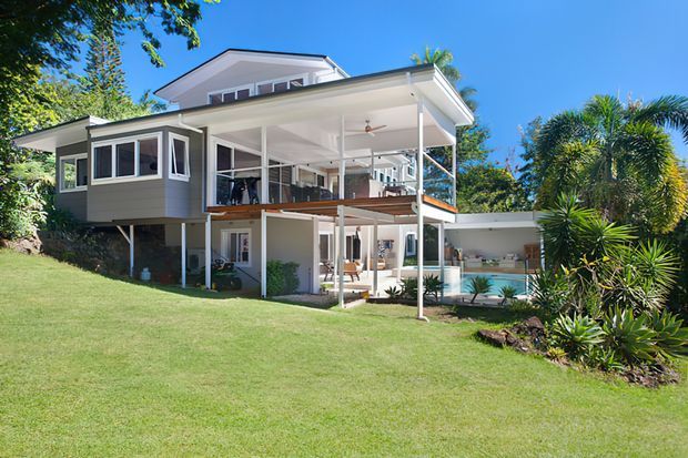 A bright exterior view of a multi-level contemporary house set within a landscaped garden. The home features pale horizontal cladding, broad roof overhangs, extensive covered outdoor living areas, white structural posts and balustrades, timber decking, large glazed openings, and a pool or water feature beside the lower level. Tropical planting and a gently sloping lawn surround the elevated structure.