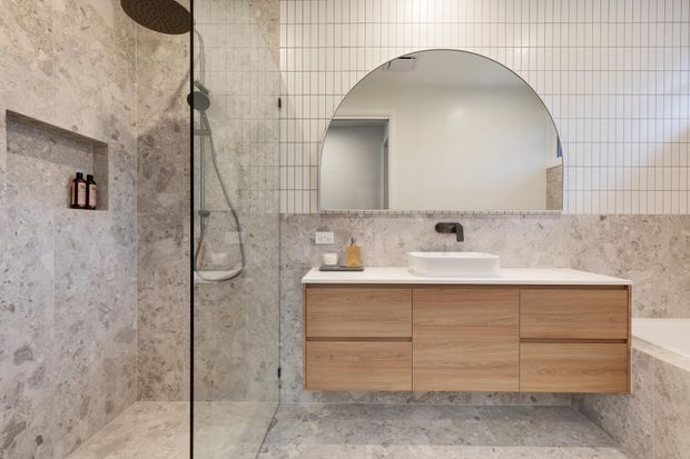 Contemporary bathroom featuring a floating timber vanity with an integrated white basin, wall-mounted black tapware, a large arched mirror, pale stone wall and floor finishes, and a glass-enclosed shower with matching stone cladding. A freestanding or built-in bath is partially visible at right.