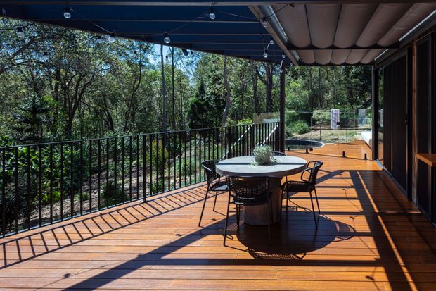 A covered outdoor timber deck overlooking dense native woodland. The deck features a round dining table with four chairs, black metal balustrading, overhead string lights, and a curved padded ceiling or awning. Glazed and timber-framed doors open along the right side, with a landscaped garden and a glimpse of a pool or water feature beyond.