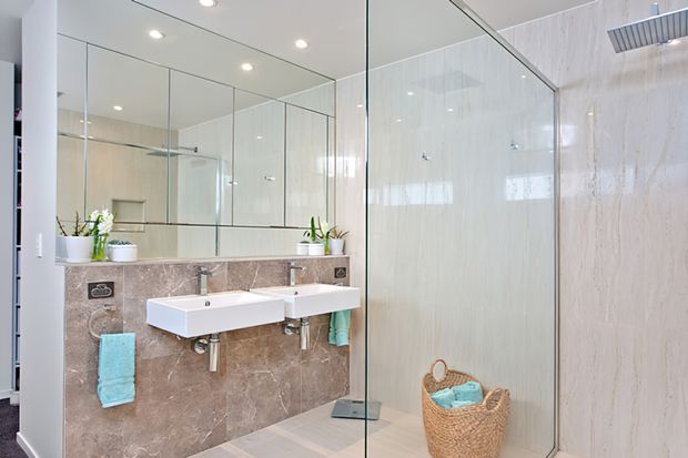 Bright contemporary bathroom with a double wall-mounted vanity, large mirrored wall, pale stone-look wall panels, glass shower enclosure, recessed ceiling lights, and woven storage baskets with turquoise towels.