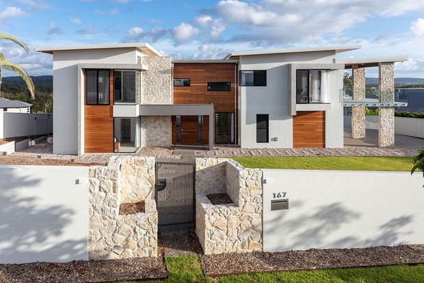 Front exterior view of a contemporary multi-level residence with a flat-roofed composition, crisp white rendered walls, warm timber-clad sections, and prominent light natural-stone feature walls and columns. The home includes projecting box forms, extensive glazing, a covered entry, landscaped frontage, and a white boundary wall with stone gate piers. A distant water or coastal landscape is visible beyond the property.