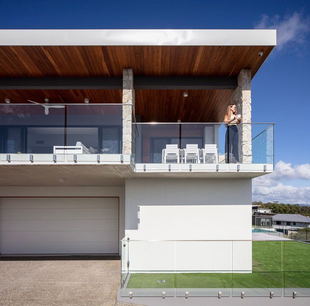 Contemporary multi-level residential home exterior photographed from the street, featuring a white rendered facade, large glass balcony balustrades, stone-clad structural columns, a timber-lined covered upper terrace, and an integrated garage. Outdoor dining furniture is visible on the upper balcony, with a small landscaped lawn and neighbouring homes beyond.