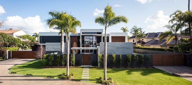 Street-facing view of a contemporary multi-level residence with a strongly horizontal composition, flat rooflines, dark and light rendered facade sections, timber-look entry and garage doors, broad glazing, and a low masonry privacy wall. A manicured lawn, stepping-stone path, palms, and adjoining driveways frame the suburban property.
