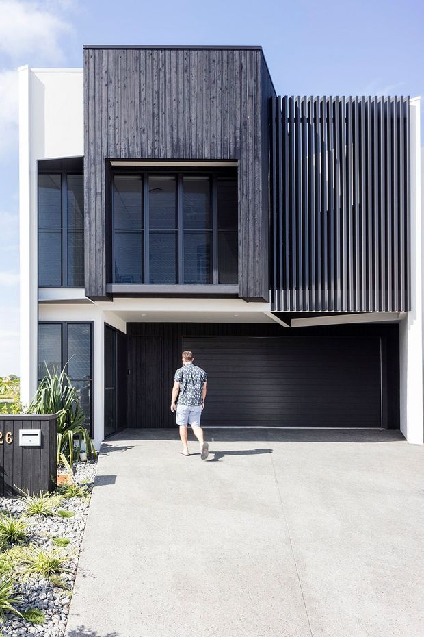 Street-facing view of a contemporary multi-storey townhouse facade with a dark vertical timber-clad upper volume, projecting vertical fins, large dark-framed windows, white rendered walls, and an integrated double garage. A concrete driveway and low-maintenance gravel planting strip occupy the foreground.