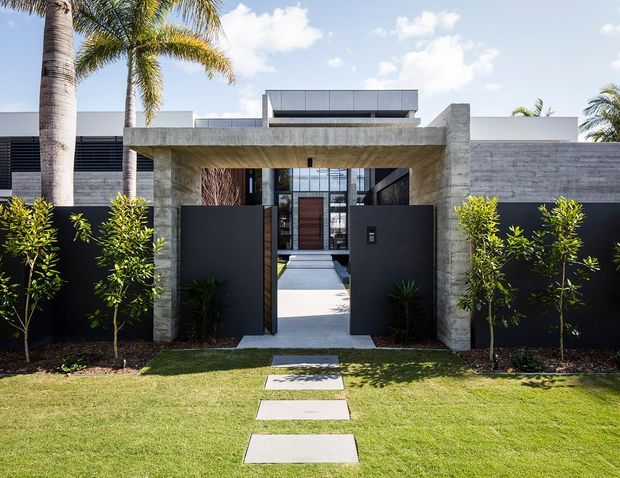 Front exterior view of a contemporary single-storey residence, focused on a wide concrete entry portal and open dark metal pedestrian gates. A paved path crosses the lawn toward a timber entry door, with full-height glazing and horizontal screening behind. The facade combines smooth white rendered volumes, dark metal panels, and textured concrete block walls, framed by palms and neatly planted hedges in bright daylight.