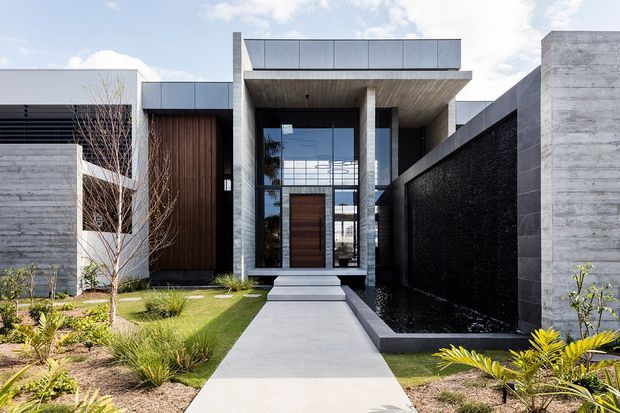 Front exterior view of a contemporary residence with a strong geometric composition, flat rooflines, projecting concrete entry canopy, dark textured masonry walls, timber-clad entry volume, and large glazed panels. A straight concrete walkway leads through a landscaped front garden to the recessed timber front door.