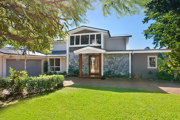 Exterior view of a contemporary multi-level residential house with a broad low-pitched roof, grey horizontal cladding, a prominent irregular-stone feature wall, timber-framed glazed entry, and upper-level clerestory-style windows. The home is set behind a manicured lawn and hedges with mature trees surrounding the frontage.