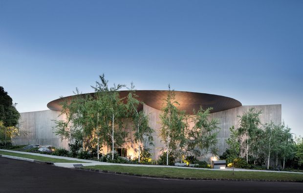 A dramatic contemporary building exterior photographed at dusk, featuring a rounded, organically shaped dark roof canopy with warm concealed lighting, pale vertical wall cladding, extensive slender trees and landscaped planting, and a curved driveway in the foreground. The low, sculptural form is set against a clear blue sky.