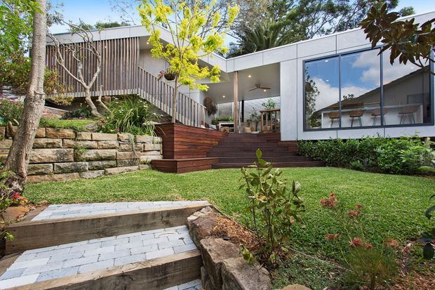A contemporary single-storey house set into a landscaped, sloping garden. The exterior combines white rendered or panelled walls, extensive dark-framed glazing, vertical timber screening, and warm timber decking and stairs. Natural stone retaining walls and paved garden steps lead up to a covered outdoor terrace and the home’s living spaces, visible through the glazing.