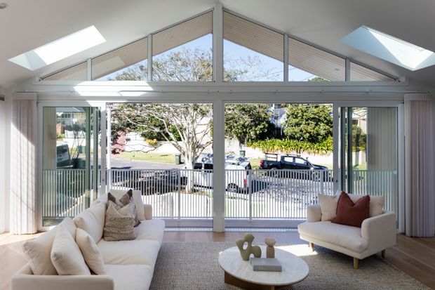 Bright contemporary living room with a high pitched glazed gable wall, extensive floor-to-ceiling windows and sliding doors opening onto a balcony or terrace. A neutral sectional sofa and armchair surround a round coffee table, while skylights, sheer curtains and light timber flooring create an airy interior.