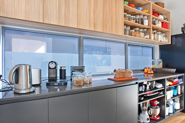 A contemporary kitchen with a long dark benchtop, matte dark lower cabinetry, extensive light timber overhead cabinetry, and open timber shelving filled with pantry jars and kitchenware. Large horizontal windows provide abundant daylight, while small appliances and baking equipment sit along the benchtop and open shelving.