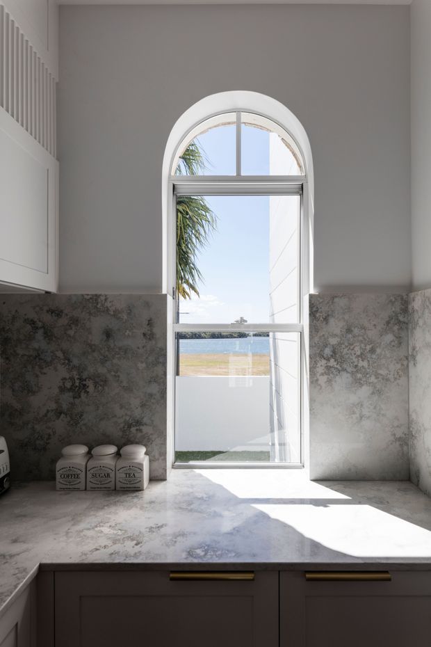 Bright kitchen work area featuring a large arched window with white framing, a mottled grey stone or stone-look splashback and benchtop, and dark grey lower cabinetry with brass pulls. Strong daylight enters through the window, with a glimpse of water and outdoor landscape beyond.