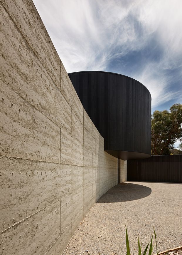 Exterior architectural view of a sculptural building composed of a tall rough concrete wall and a dark, vertically ribbed curved volume projecting above a gravel forecourt. The dramatic cantilevered form creates a deep shadowed recess, with sparse planting and trees visible at the perimeter under a partly cloudy sky.